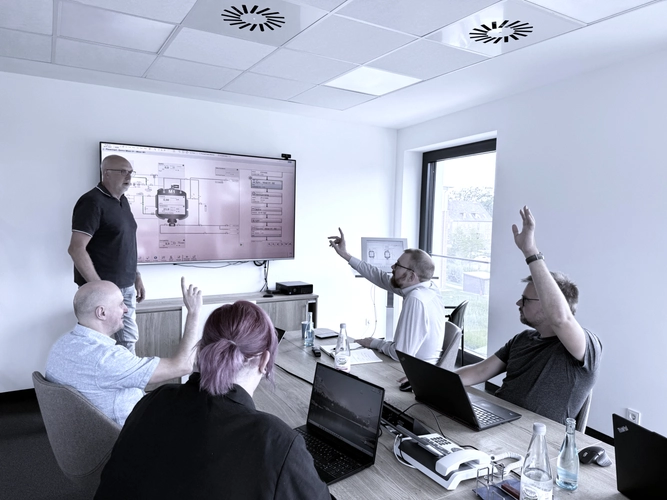 Eine Gruppe von Personen in einem Besprechungsraum, einige heben die Hand, während jemand an einem Bildschirm präsentiert.